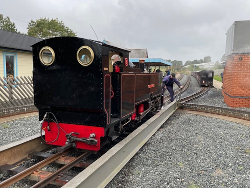 The black steam engine sitting on a turntable. At the far end is the driver, pushing against one of the handles to turn the train. Beyond the turntable are three tracks and the rest of the station.